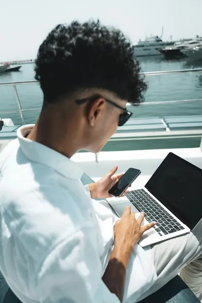 A boater aboard a powerboat in a marina is using his digital devices: a laptop and a smartphone